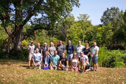 Two rows of people pose in a lawn outside. Behind them are trees and shrubs that surround a pond.