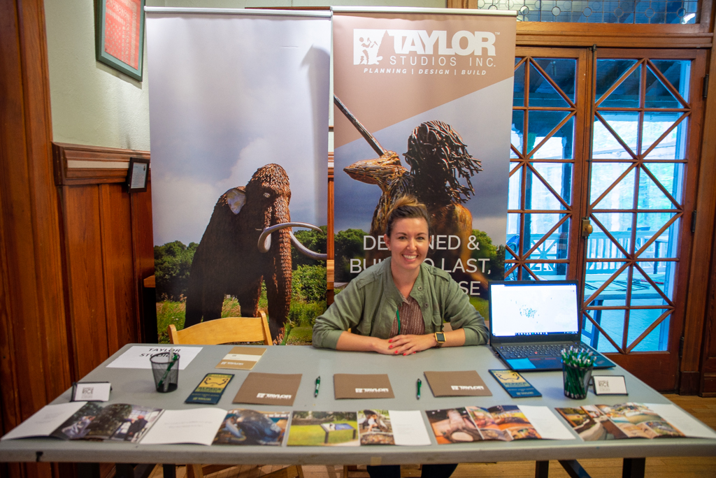 A woman smiles and sits at a folding table with an assortment of pamphlets. Behind her is a banner of a wooly mammoth and a long-haired person with a spear.