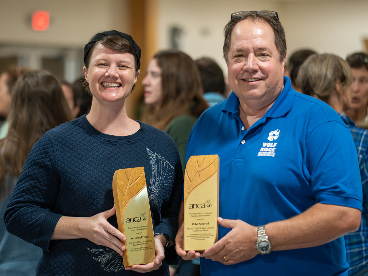 Two people stand and pose in a crowded room. The two people each hold a small rectangular wooden item with a plaque on it.