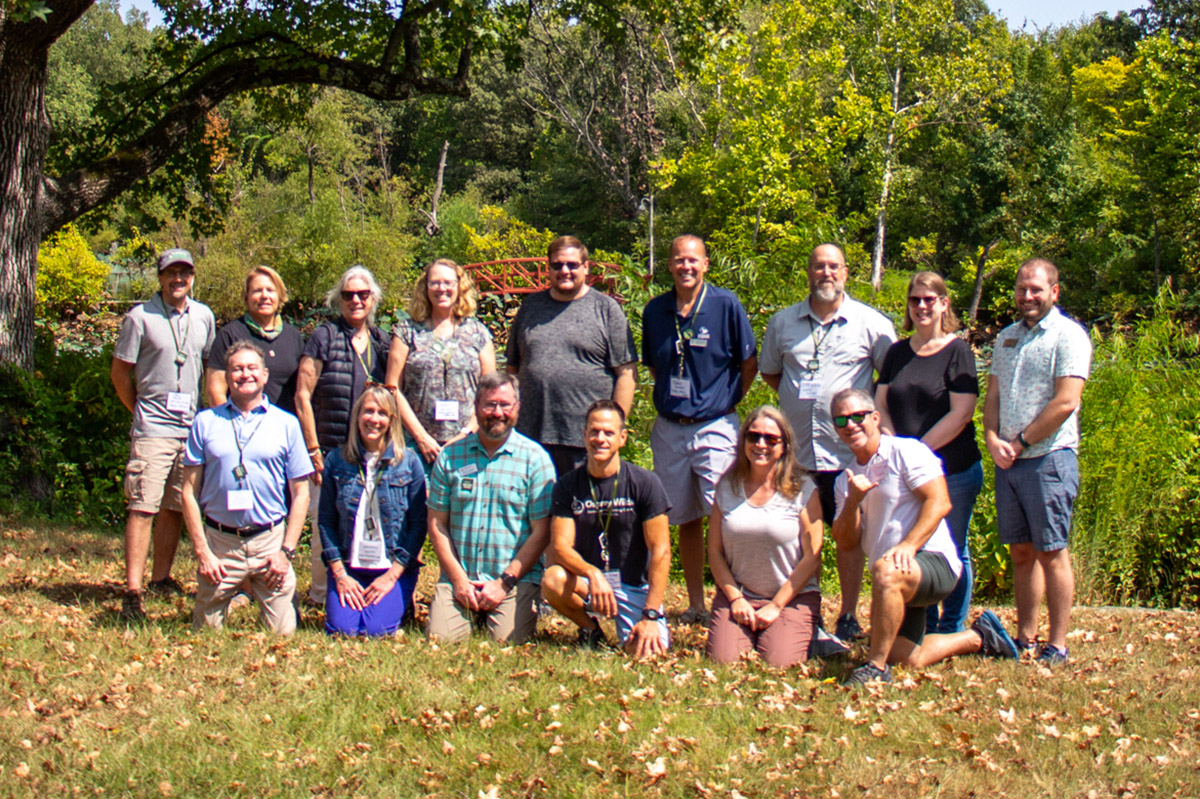 Two rows of people pose on a lawn. Behind them are large trees and shrubs that surround a pond.