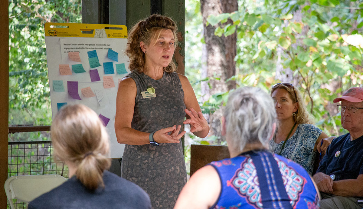 A person stands and speaks in front of a group, gesturing with their hands. Behind them, green foliage.