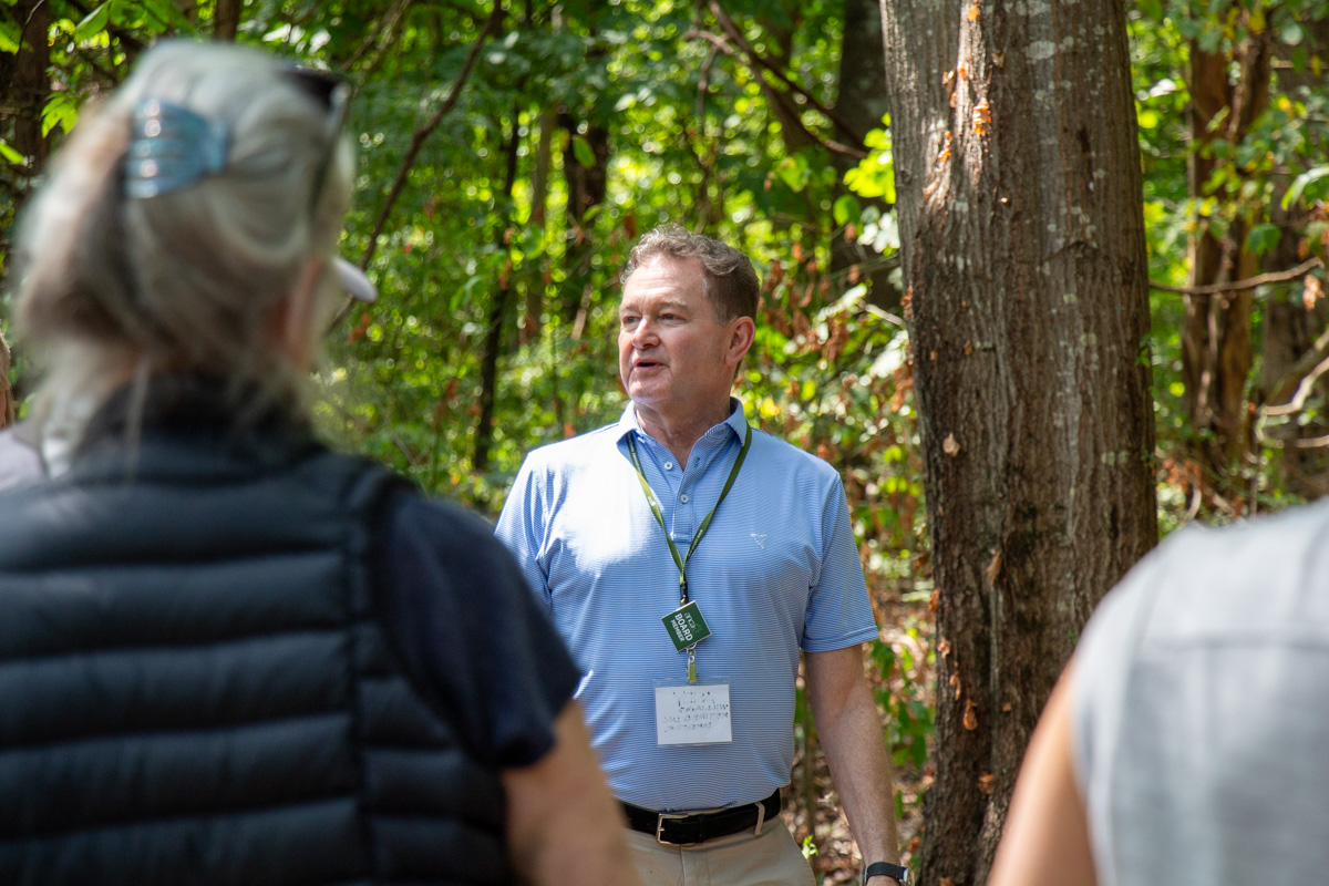 A light-skinned person with short hair wears a light-blue polo shirt in a forest. They speak to a group of people in the foreground.