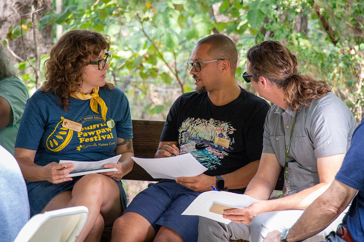 Three people sit on a bench outside. Each person holds a sheet of paper, and the three lean in, engaged in conversation. In the background, green foliage.