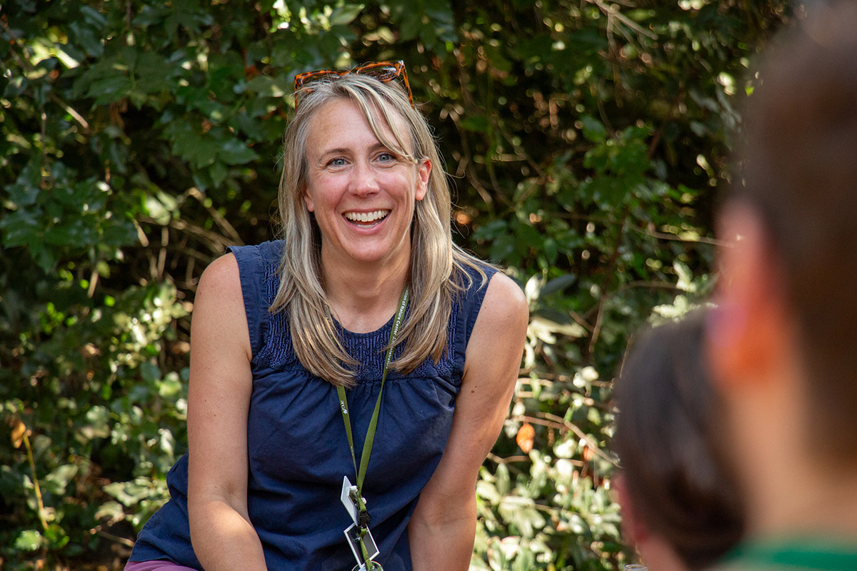 A long-haired person smiles widely at two people in the foreground. Behind them, foliage speckled with sunlight.
