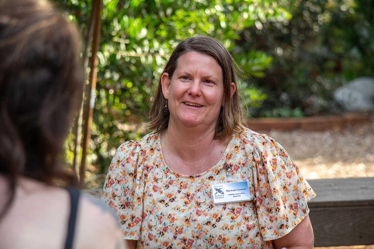 A light-skinned person in a floral shirt smiles at a person in the foreground. Sunlit foliage stands behind them.