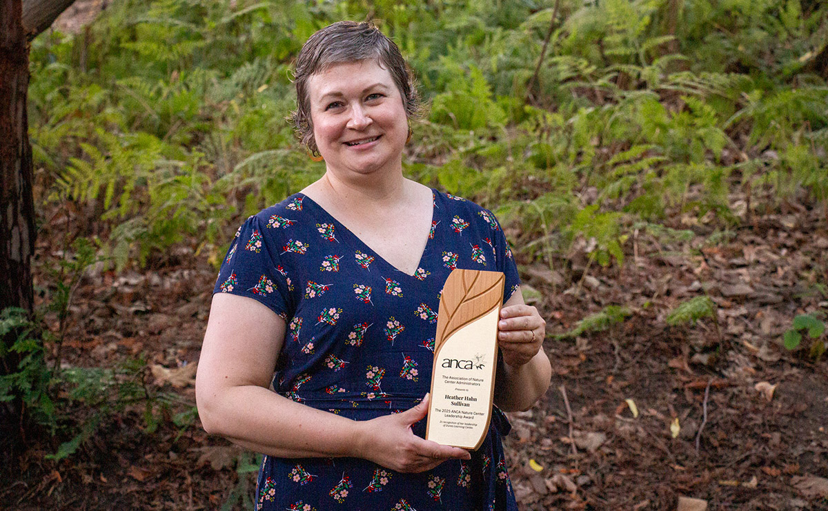 A short-haired person in a blue patterned dress poses, holding a wooden and metal plaque. Behind the person is a hillside of ferns.