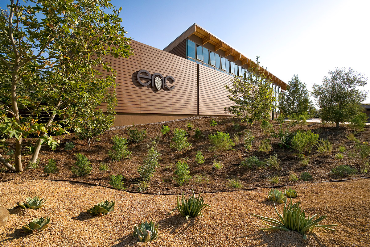 A sandy garden with sparse succulents stands before a long building with horizontal wood siding. The letters "E-N-C" sit on one section of the siding. Above, blue sky and sunshine.