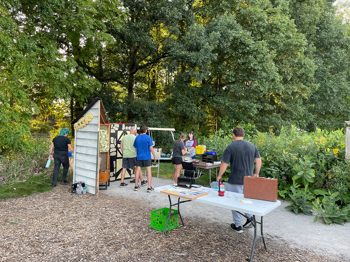 A station of folding tables is set up outdoors on a path. A group of people stand around the tables.