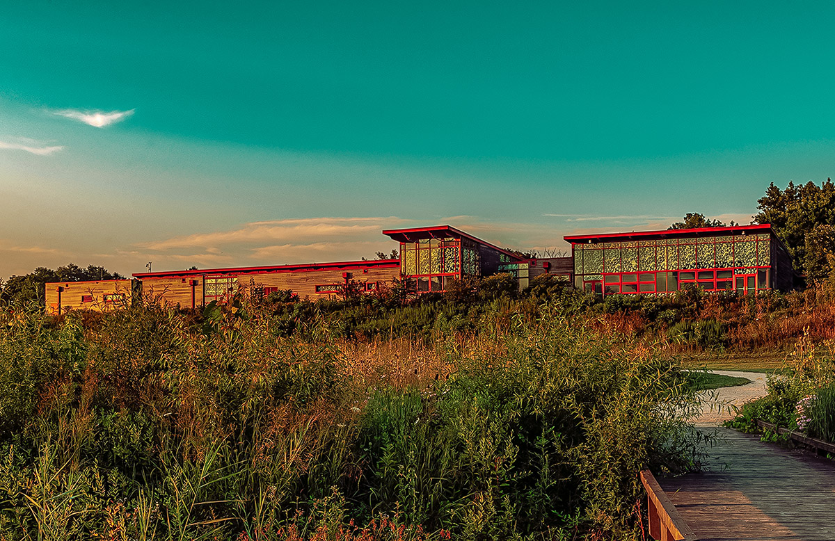 A walkway and winding path lead up for a long building surrounded by prairie. Above, blue sky.