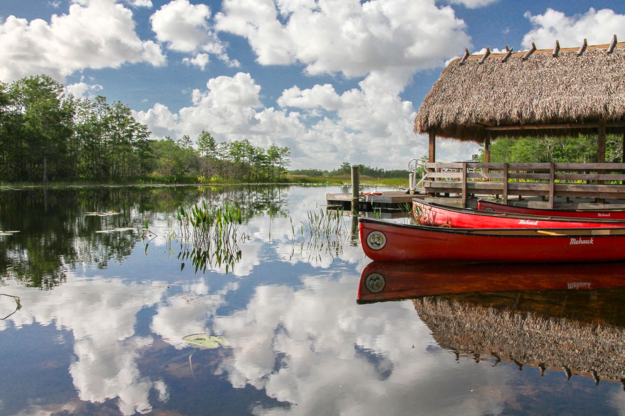 Red canoes sit on the water by a simple dock. Puffy clouds above reflect on the still water.