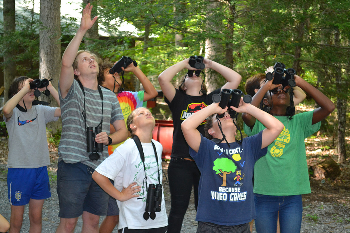 A group of seven children and a young adult stand in the forest. Most of the children look upward through binoculars, while the young adult raises their hand upward in explanation.