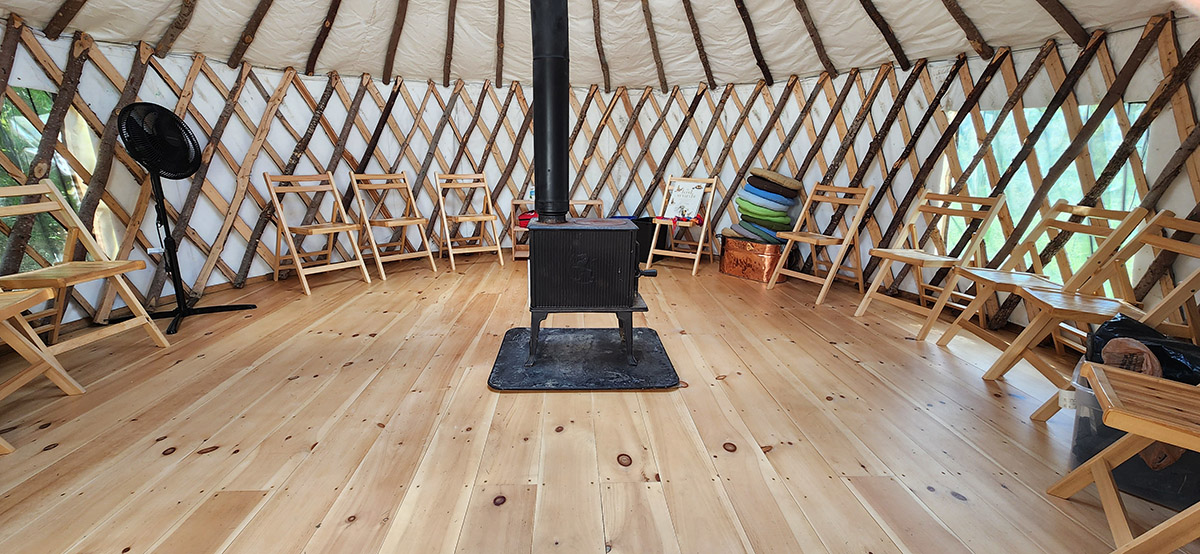 An inside view of a yurt: a black wood stove stands in the middle, folding wooden chairs align the outer perimeter of the room.