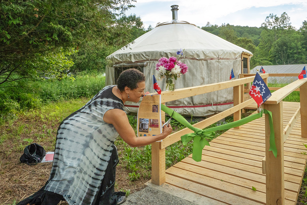 A dark-skinned person with short hair and a long black-and-white dress holds scissors over a green ribbon in front a wooden walkway that leads to a yurt.