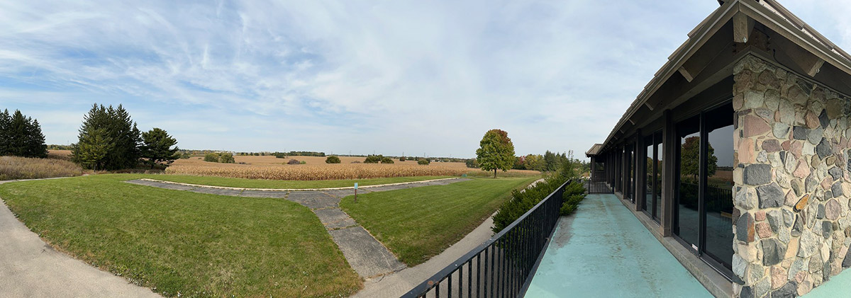 A panoramic view. A stone building with glass windows stands to the right. To the left, a mowed area with cornfields beyond.