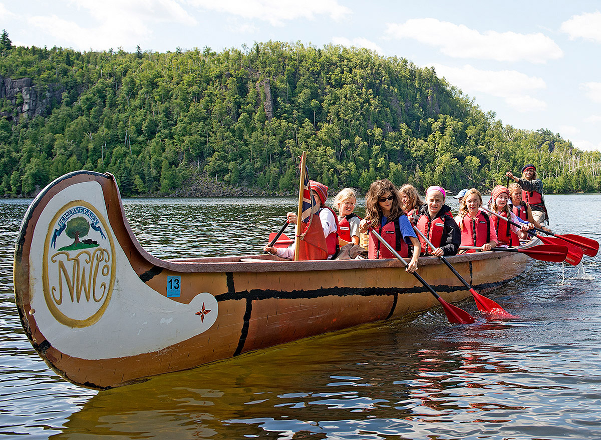 A group of children in a canoe on the water. In the background, a forested hill and blue sky with clouds.