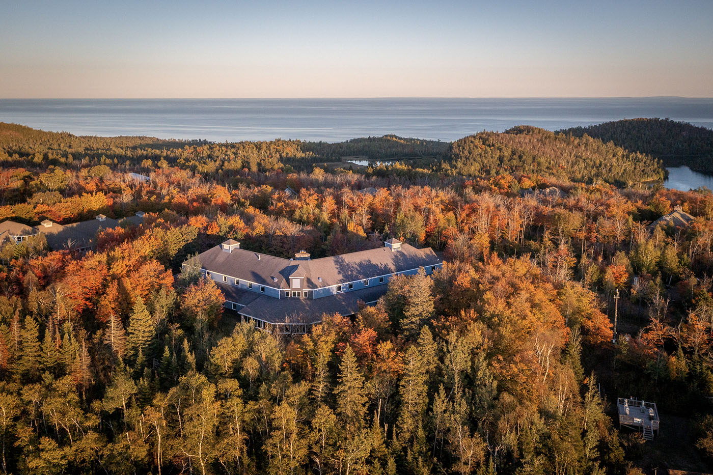 An aerial view of the MAC Lodge at Wolf Ridge Environmental Learning Center. A building is surrounded by green, yellow, and orange trees in an evening sunlight. A lake's edge is in the background.