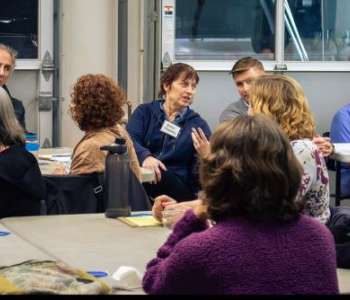 A group of people sit at tables indoors. One person gestures with their hand and speaks, others look and listen. A group of people sit at tables indoors. One person gestures with their hand and speaks, others look and listen.