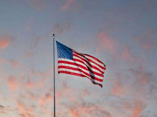 An American flag waves against a background of a sunset. Whispy pink clouds stand out against a darkening blue sky.
