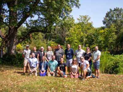 Two rows of people pose in a lawn outside. Behind them are trees and shrubs that surround a pond.