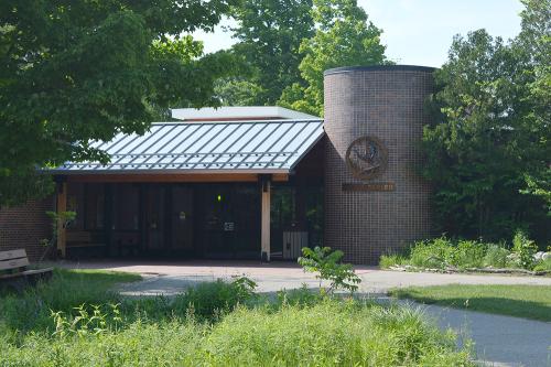 A one-story building with a slanted roof over an entryway, surrounded by trees. On the right side is a large cylindrical feature. In front, paved trails lead to the building.
