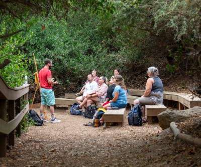 A group of people sit on benches outside, facing a person standing. A group of people sit on benches outside, facing a person standing.