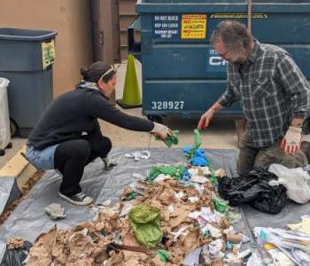 Two people sort through a pile of trash by a dumpster.