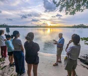 A group of people stand beside a body of water at sunset. One person speaks and gesticulates.